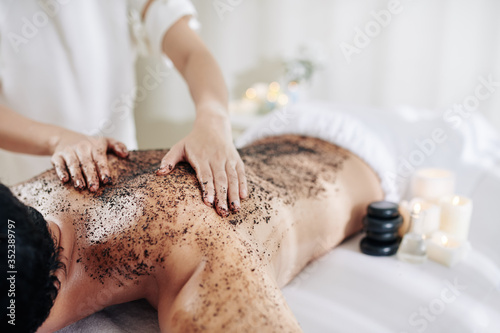Cosmetologist applying coffee peeling on back of male client to exfoliate ot before massage