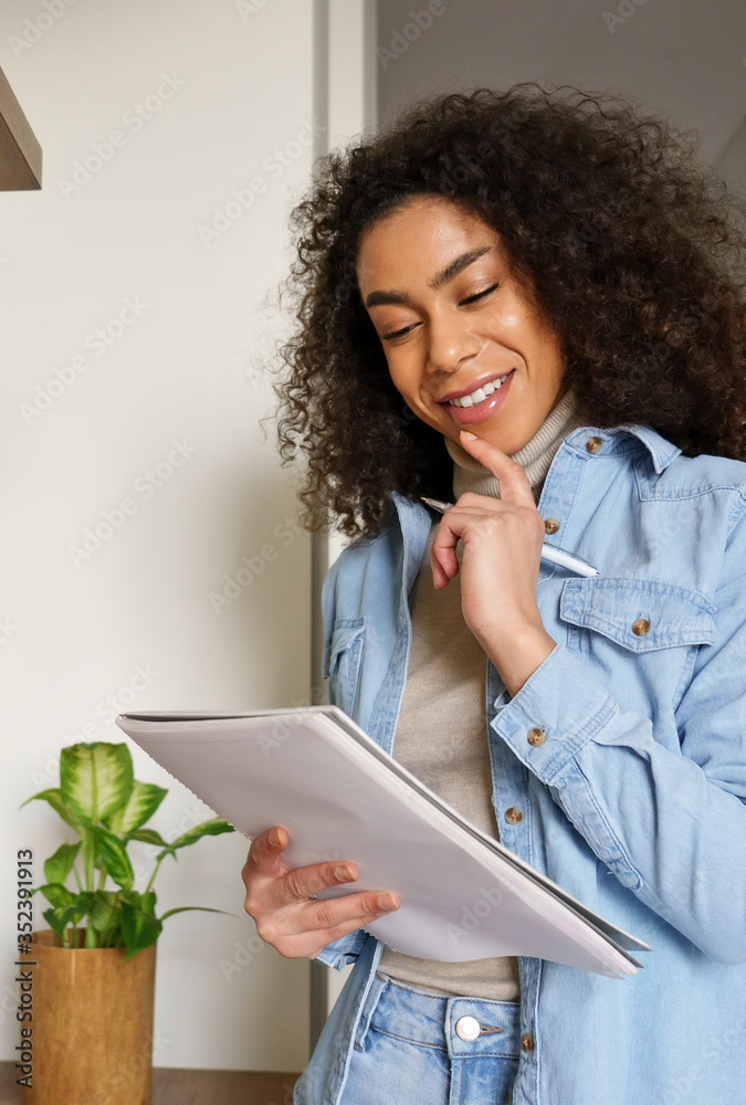 Smiling african american teen girl college student, remote worker, writer study work from home office thinking on writing creative essay idea doing homework holding notebook reading notes. Vertical Smiling african american teen girl college student, remote worker, writer study work from home office thinking on writing creative essay idea doing homework holding notebook reading notes. Vertical