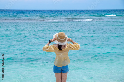 Young woman in yellow blouse and short blue jean with straw hat standing on the beach with blue sky that relaxing and enjoying on vacation.