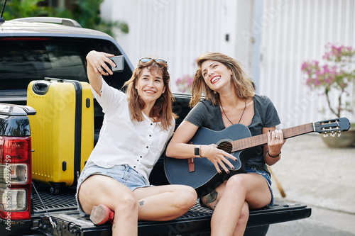 Pretty positive young woman taking selfie with her friend playing guitar when sitting in car trunk