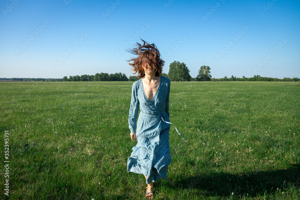 A cheerful dark-haired woman smiles, walks along the green field dressed in a romantic blue dress on the smell, wicker bag with a bouquet  on a summer day. 