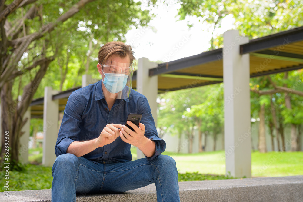 Young man using phone with mask and face shield while sitting at the park - obrazy, fototapety, plakaty 