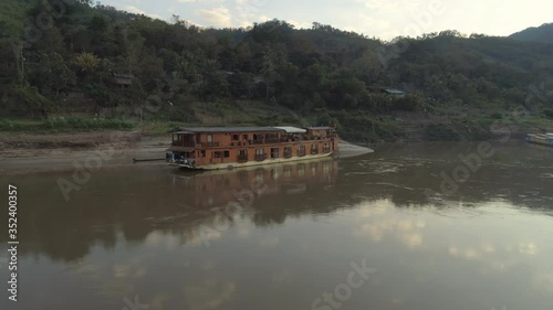 Aerial shot of cruise ship moored at riverbank against forest, drone panning over river against mountains during sunset - Mekong River, Laos photo