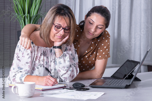 Smiling mature woman writing on a notebook surrounded by a laptop and a tablet while being embraced by her daughter. Home office and family concept.