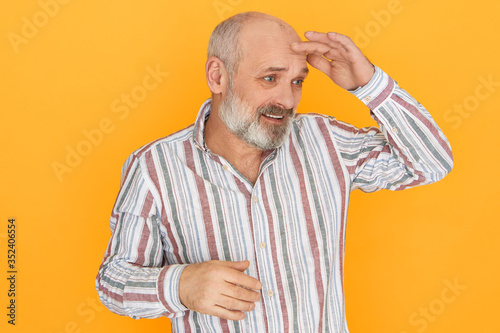 Studio image of frustrated puzzled senior bald unshaven man holding palm on forehead to protect eyes from sun, looking into dustance or searching for something. Human facial expressions photo