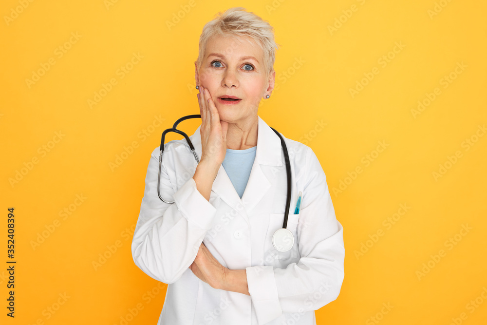 Human facial expressions, emotions and feelings. Studio image of emotional surprised retired woman practitioner holding hand on cheek and opening mouth, shocked with anamnesis or diagnosis - obrazy, fototapety, plakaty 