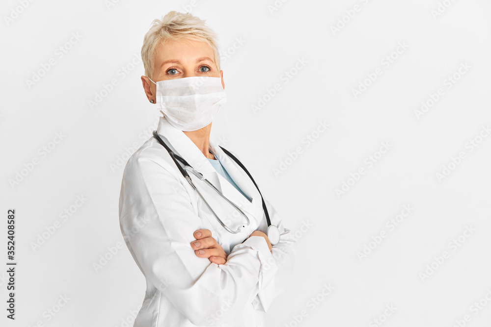 Confident mature woman physician in protective medical mask and uniform crossing arms on her chest posing against blank studio wall background with copy space for your advertising information Confident mature woman physician in protective medical mask and uniform crossing arms on her chest posing against blank studio wall background with copy space for your advertising information