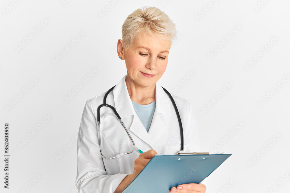 Studio shot of attractive middle aged blonde woman physician with stethoscope around her neck posing isolated with pen and clipboard, making medical records, prescribing treatment for patient Studio shot of attractive middle aged blonde woman physician with stethoscope around her neck posing isolated with pen and clipboard, making medical records, prescribing treatment for patient