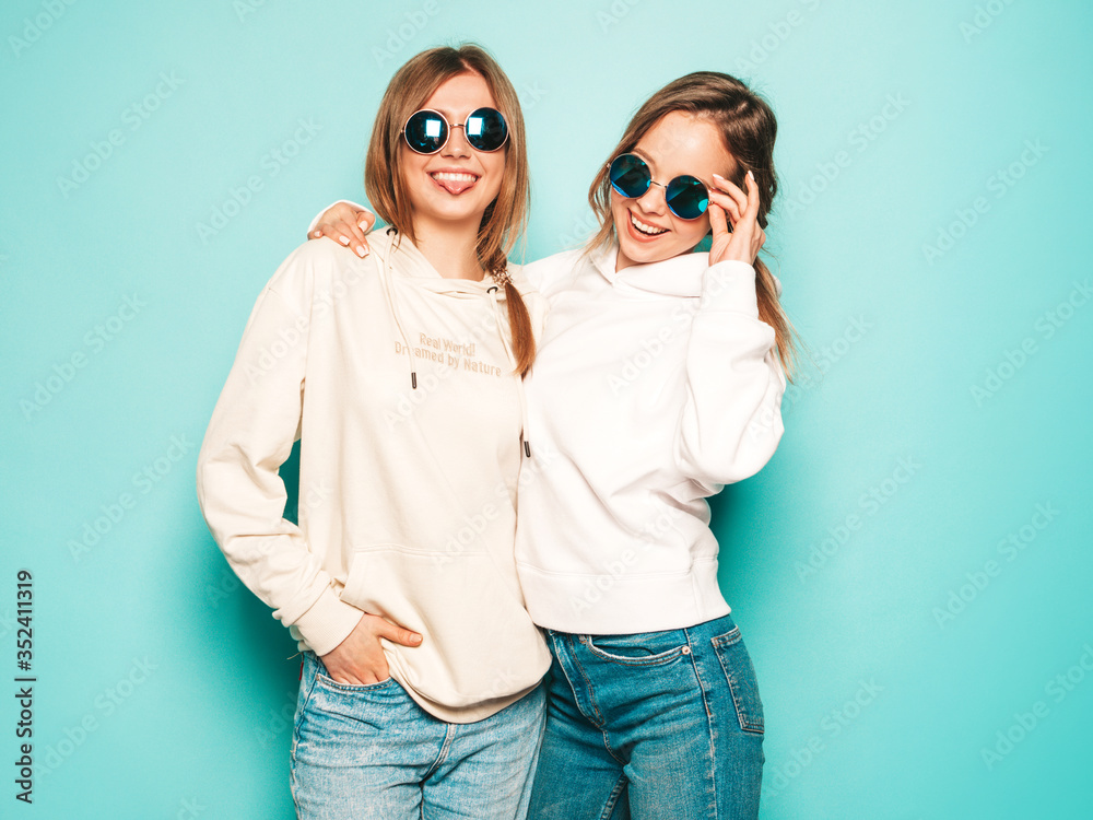 Two young beautiful blond smiling hipster girls in trendy summer hoodie clothes. Sexy carefree women posing near blue wall. Trendy and positive models having fun in sunglasses