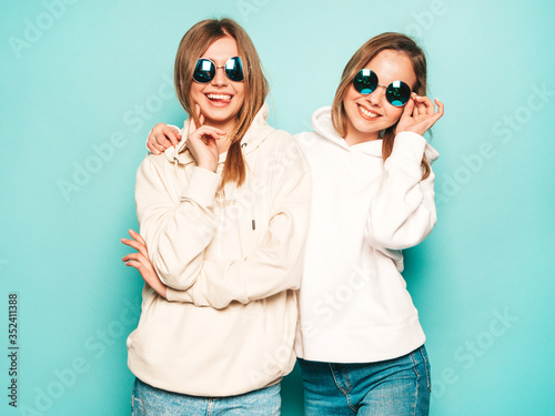 Two young beautiful blond smiling hipster girls in trendy summer hoodie clothes. Sexy carefree women posing near blue wall. Trendy and positive models having fun in sunglasses © halayalex