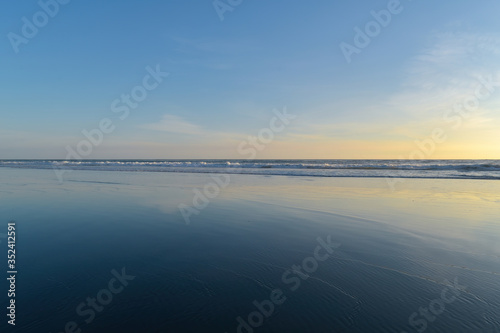 Tasman Sea horizon and faint clouds reflecting in thin water film over black sand in evening light