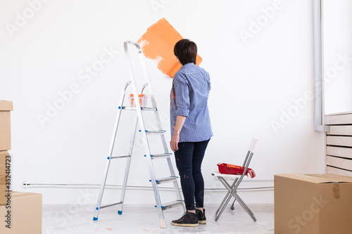 Happy middle-aged woman painting interior wall with paint roller in new house. A woman with roller applying paint on a wall.