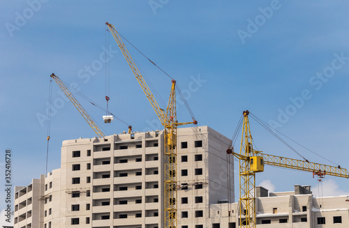 Construction site. Unfinished apartment buildings. Construction workers laying bricks on top. Special industrial cranes in the middle. Blue sky background.