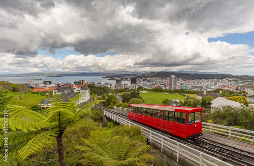 Red cable car moving up the mountain in the foreground and panorama of Wellington in the background. Beautiful blue sky covered with clouds. 