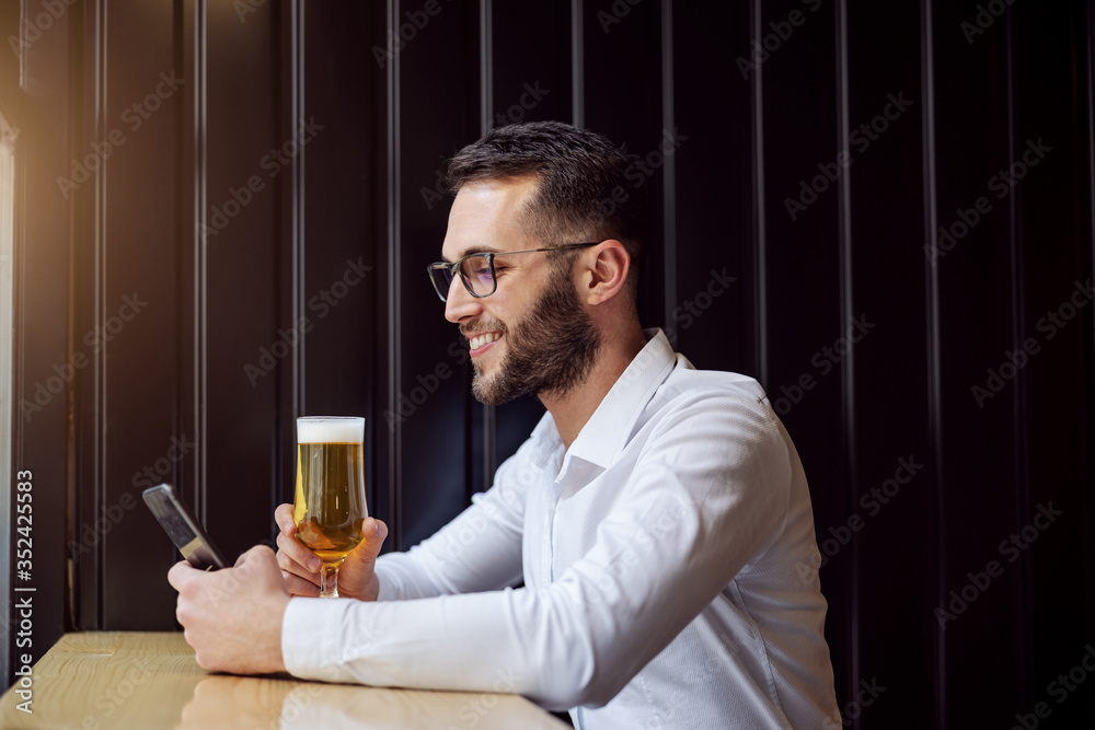 Side view of happy businessman sitting in pub, holding glass of beer and using smart phone for internet surf. After work activities.