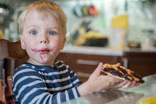 Little cute blond boy bully without permission of his mother broke off piece of berry pie and eats it. Surprise of baby