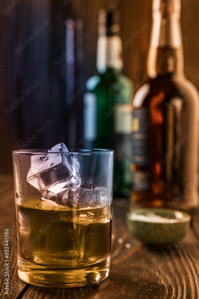 A glass of whiskey with ice, in the background are bottles on a wooden table of a bar counter, shallow depth of field, selective focus. The concept of alcoholic drinks in a roadside bar. A glass of whiskey with ice, in the background are bottles on a wooden table of a bar counter, shallow depth of field, selective focus. The concept of alcoholic drinks in a roadside bar.