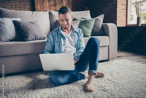 Photo of handsome homey guy relaxing sitting comfy fluffy carpet near couch browsing notebook freelancer remote work stay home good mood quarantine time weekend living room indoors