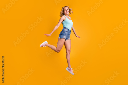 Full size profile photo of attractive pretty lady good mood jump high up excited expression walking down street wear teal tank-top denim shorts shoes isolated yellow color background