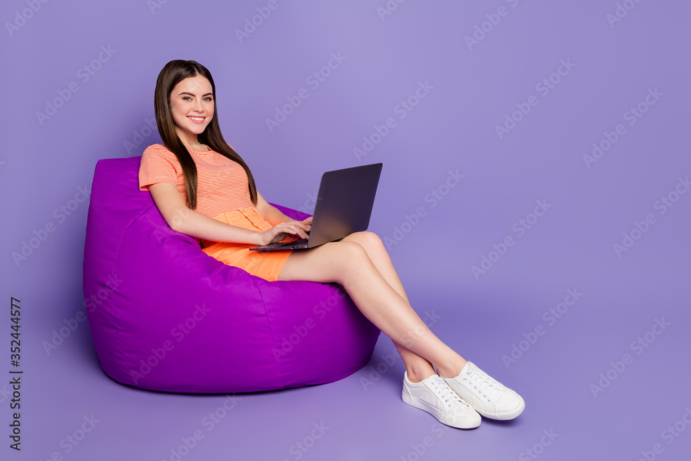 Full size profile photo of cheerful lady sitting comfy beanbag browsing notebook freelance work wear orange striped t-shirt jeans mini skirt isolated pastel purple color background Full size profile photo of cheerful lady sitting comfy beanbag browsing notebook freelance work wear orange striped t-shirt jeans mini skirt isolated pastel purple color background