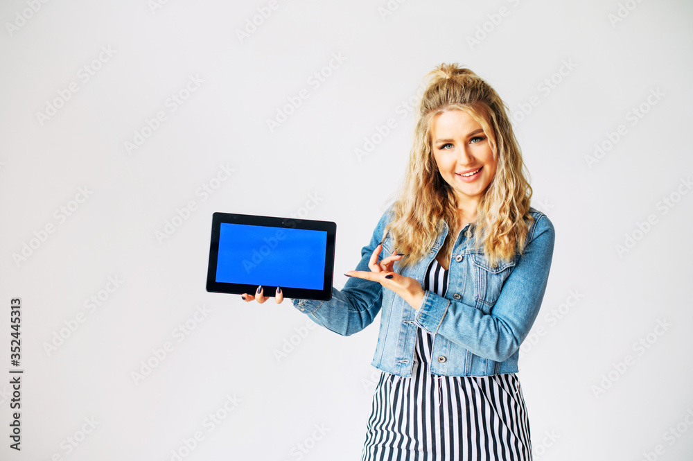 Look at this. Beautiful young woman with a tablet computer in hands stands on a white background. Woman points finger to blank tablet screen, empty space for inserting pictures, text