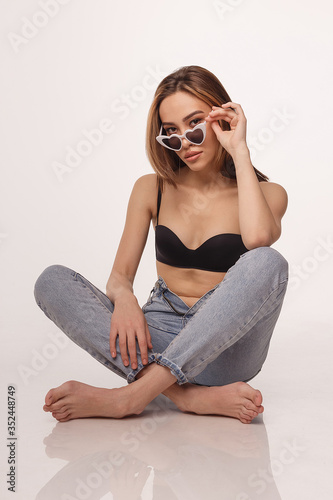 portrait of sexy asian woman with long hair posing in black lingerie, sunglasses, jeans on white studio background with bare feet. model tests of skinny girl in bra. attractive female sitting on floor