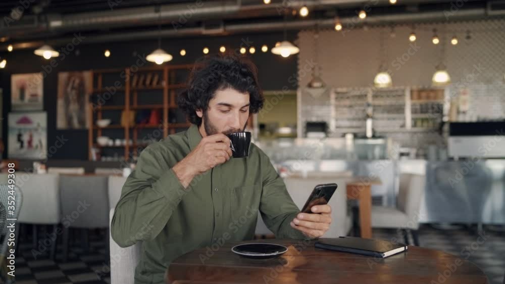 Smiling young caucasian man in cafe drinking coffee while texting on smartphone in cafe
