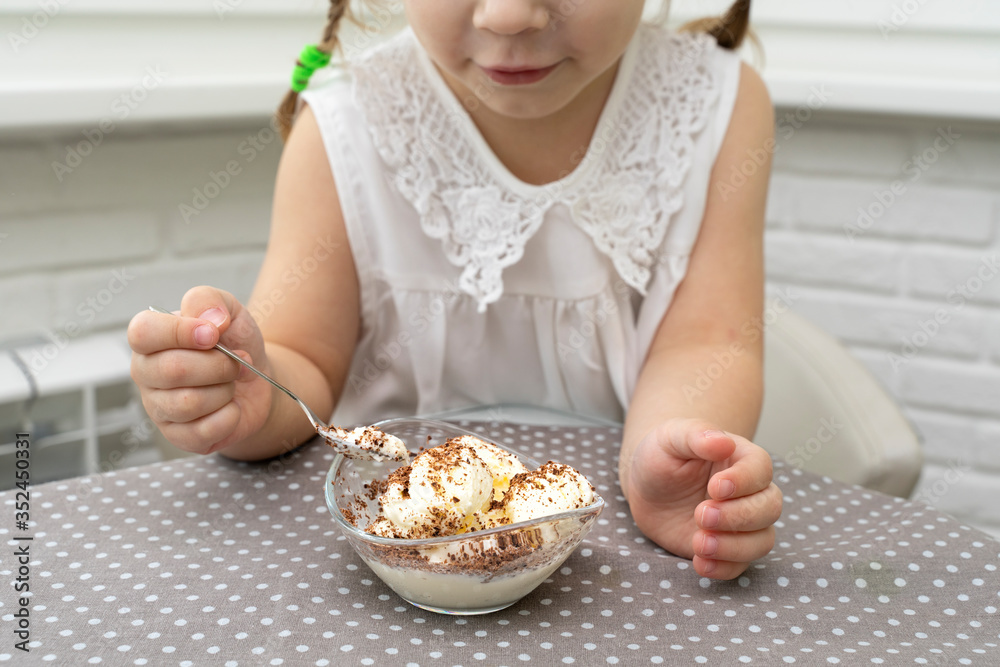 little girl in a white blouse happily eats ice cream from a snail at a table in the kitchen