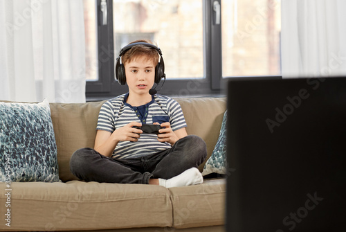leisure, technology and children concept - smiling boy in headphones with microphone and gamepad playing video game at home