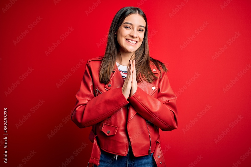 Young beautiful brunette woman wearing casual jacket standing over red background praying with hands together asking for forgiveness smiling confident. Young beautiful brunette woman wearing casual jacket standing over red background praying with hands together asking for forgiveness smiling confident.