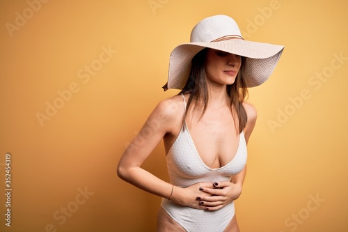 Young beautiful brunette woman on vacation wearing swimsuit and summer hat with hand on stomach because indigestion  painful illness feeling unwell. Ache concept.