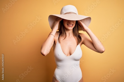Young beautiful brunette woman on vacation wearing swimsuit and summer hat with hand on headache because stress. Suffering migraine.