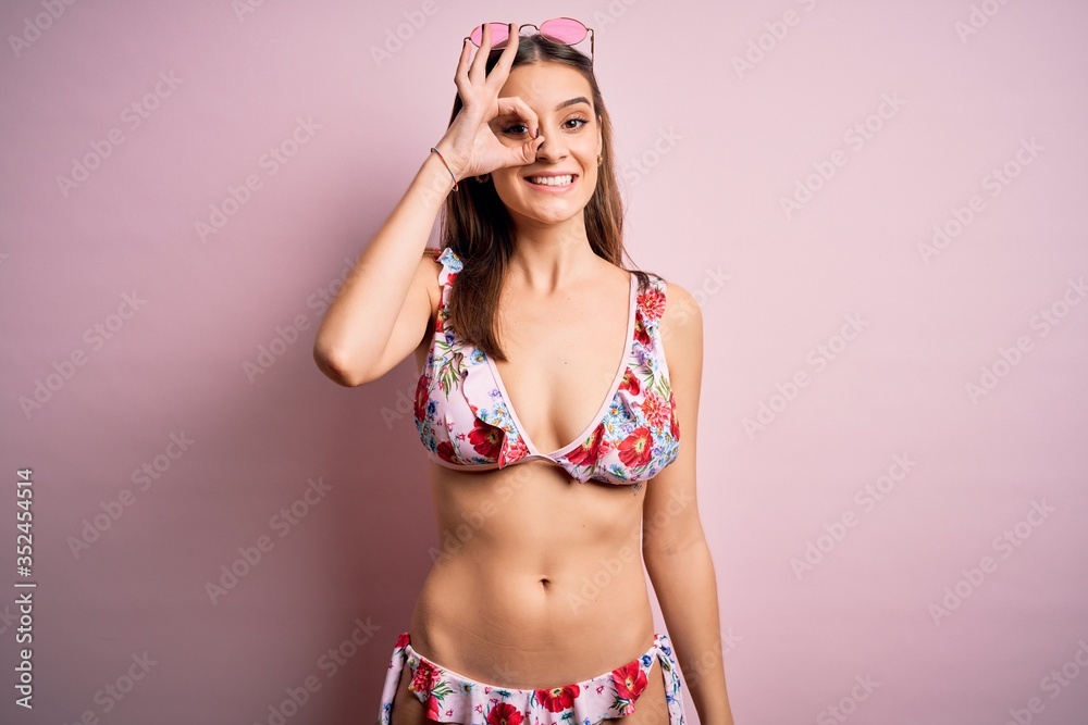 Young beautiful woman on vacation wearing bikini and sunglasses over pink background doing ok gesture with hand smiling, eye looking through fingers with happy face. Young beautiful woman on vacation wearing bikini and sunglasses over pink background doing ok gesture with hand smiling, eye looking through fingers with happy face.