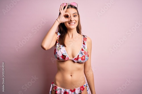 Young beautiful woman on vacation wearing bikini and sunglasses over pink background doing ok gesture with hand smiling, eye looking through fingers with happy face. © Krakenimages.com