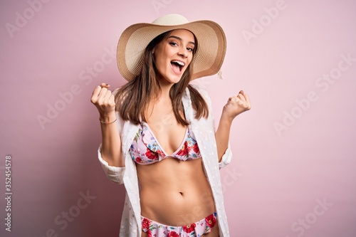 Young beautiful woman on vacation wearing bikini and summer hat over pink background celebrating surprised and amazed for success with arms raised and open eyes. Winner concept.