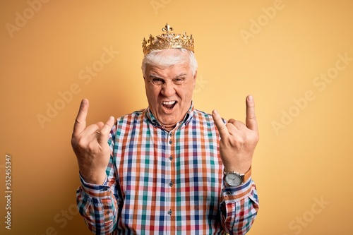 Senior handsome hoary man wearing golden crown of king over isolated yellow background shouting with crazy expression doing rock symbol with hands up. Music star. Heavy concept.