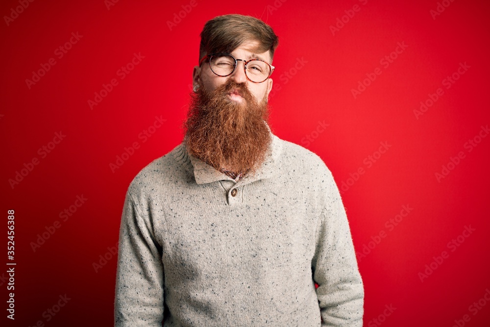 Handsome Irish redhead man with beard wearing casual sweater and glasses over red background looking at the camera blowing a kiss on air being lovely and sexy. Love expression.