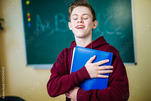 Student of higer school is standing in dark hoodie near the green blackboard in the classroom. Hugging book . Front view. School education concept. photo