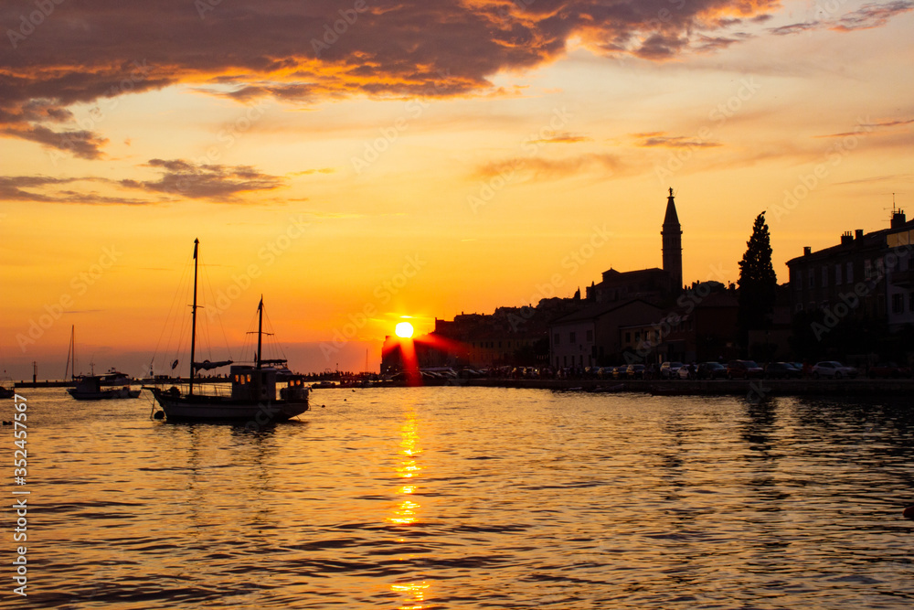 Sunset in Rovinj, Croatia, with a boat sailing in the Adriatic Sea and the sunrays reflected on the water, with the old town at the background