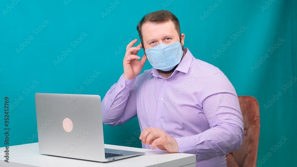 Portrait of a bearded man wearing a protective medical mask at a laptop. Male looking at the camera
