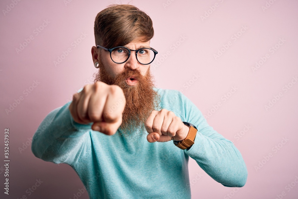 Handsome Irish redhead man with beard wearing glasses over pink isolated background Punching fist to fight, aggressive and angry attack, threat and violence