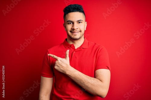Young handsome man wearing red casual polo standing over isolated background cheerful with a smile of face pointing with hand and finger up to the side with happy and natural expression on face