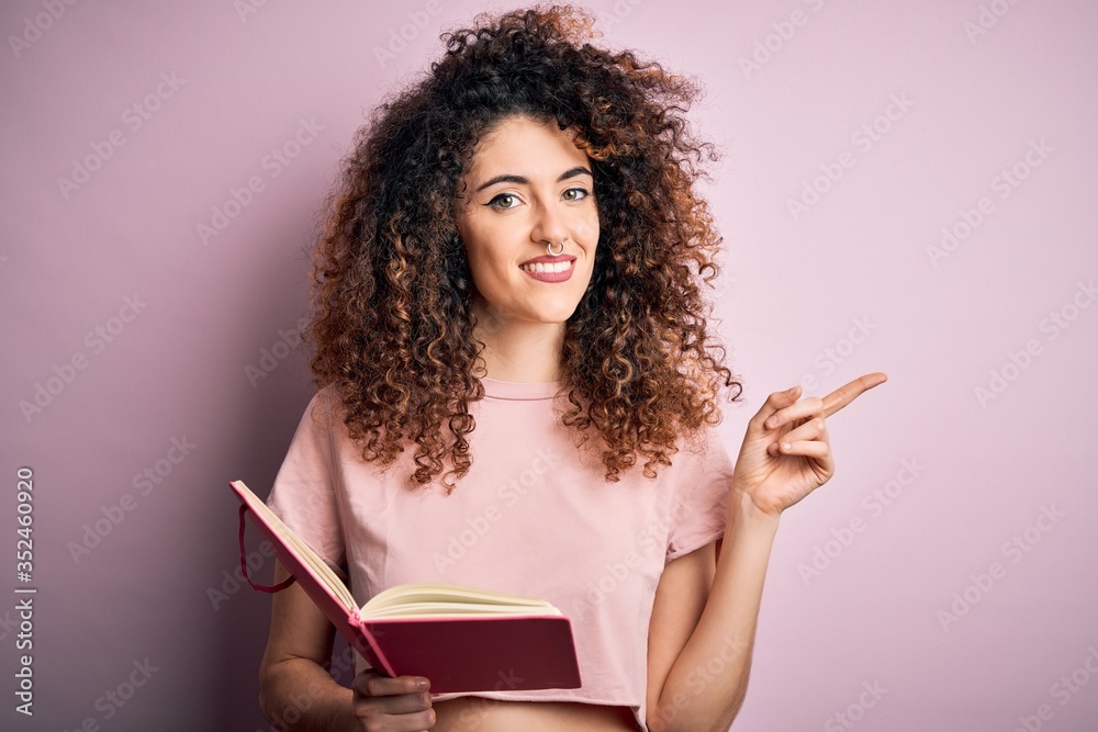 Young beautiful student woman with curly hair and piercing reading book over pink background very happy pointing with hand and finger to the side Young beautiful student woman with curly hair and piercing reading book over pink background very happy pointing with hand and finger to the side