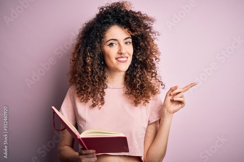 Young beautiful student woman with curly hair and piercing reading book over pink background very happy pointing with hand and finger to the side