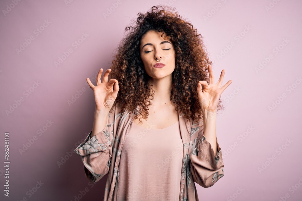 Young beautiful woman with curly hair and piercing wearing casual pink t-shirt relax and smiling with eyes closed doing meditation gesture with fingers. Yoga concept. Young beautiful woman with curly hair and piercing wearing casual pink t-shirt relax and smiling with eyes closed doing meditation gesture with fingers. Yoga concept.