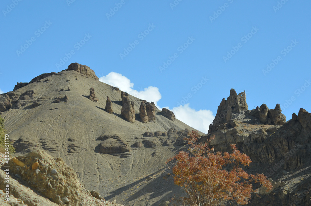 Rocky mountains in Ladakh, India are covered with unusual rock formations. On one outcrop stands a ruined stone building. The sky is blue with white clouds, and an autumn tree is in the foreground.