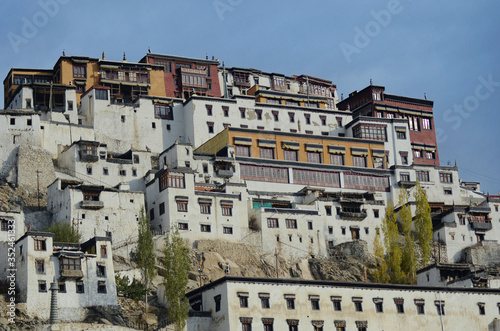 Weathered white, yellow and red buildings on a mountainside comprise part of the Thiksey Gompa near Leh in Ladakh, India. The sky is blue.