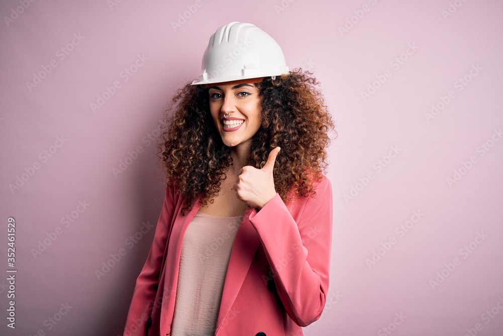 Young beautiful architect woman with curly hair wearing safety helmet over pink background doing happy thumbs up gesture with hand. Approving expression looking at the camera showing success. Young beautiful architect woman with curly hair wearing safety helmet over pink background doing happy thumbs up gesture with hand. Approving expression looking at the camera showing success.