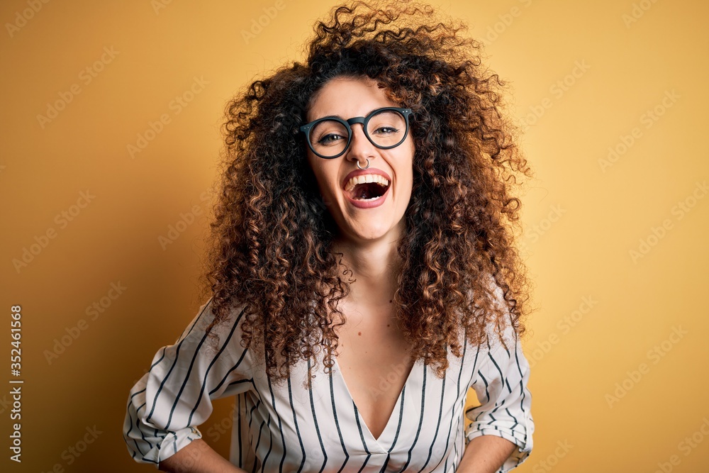 Young beautiful woman with curly hair and piercing wearing striped shirt and glasses smiling and laughing hard out loud because funny crazy joke with hands on body. Young beautiful woman with curly hair and piercing wearing striped shirt and glasses smiling and laughing hard out loud because funny crazy joke with hands on body.