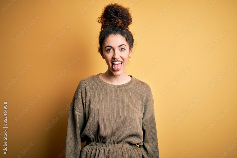 Young beautiful brunette woman with curly hair and piercing wearing casual dress sticking tongue out happy with funny expression. Emotion concept. Young beautiful brunette woman with curly hair and piercing wearing casual dress sticking tongue out happy with funny expression. Emotion concept.